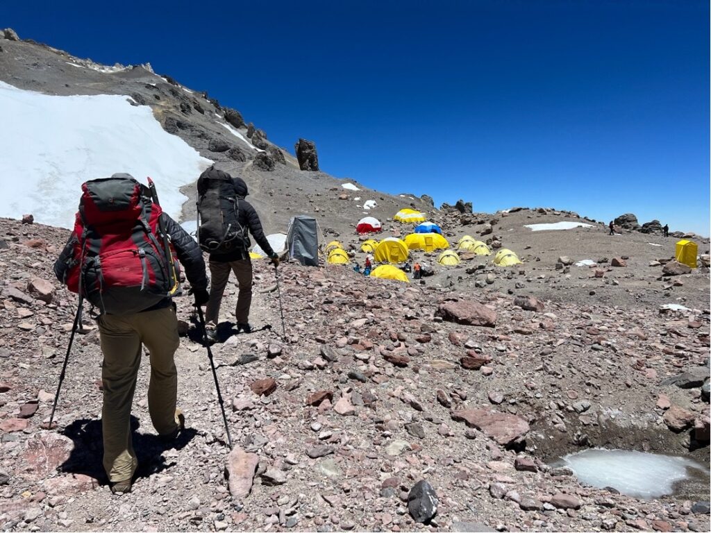 Two hikers with heavy backpacks and trekking poles walking into a camp filled with tents